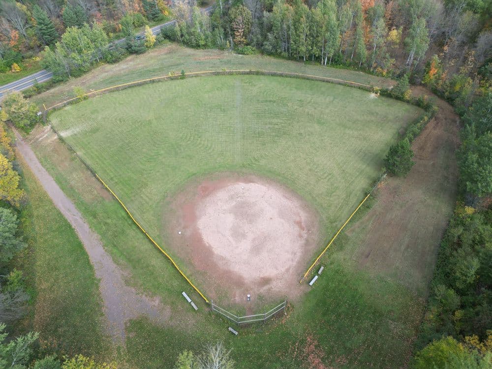 Aerial view of a baseball field surrounded by trees and a road, showcasing its green grass and infield.